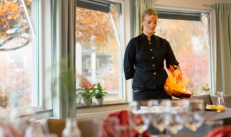 En person i svart uniform tillagar en maträtt vid bordet genom att flambera den, med lågor som stiger från pannan. Restaurangen har stora fönster där höstlöven skymtar i bakgrunden.