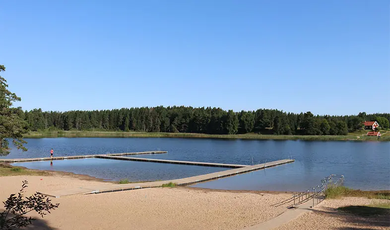 Sjö med sandstrand, bryggor och handikappramp. På andra sidan sjön finns en skog.