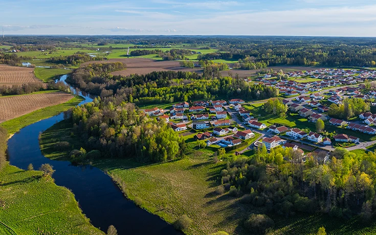 Flygfoto över landskap med slingrande å, omgivande skogspartier, åkermark och bostadsområde med vita hus. Visar en del av området där vattenrådet verkar.