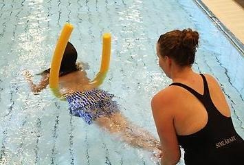 Ett barn flyter i vattnet och en simlärare håller i barnets ben.
