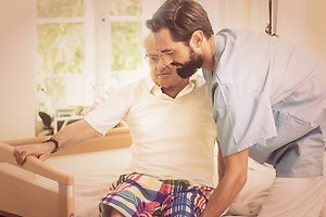 Foto på en skäggig man i vårdkläder som hjälper en äldre herre upp ur sin säng.