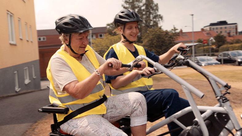 En yngre manlig sommarvikarie cyklar på en tvåsitsig cykel ihop med en äldre kvinna. Båda skrattar i solljuset.