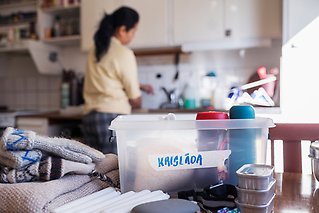 Låda med matvaror märkt "krislåda", vantar, ljus. En person står och fyller på vatten i bakgrunden.